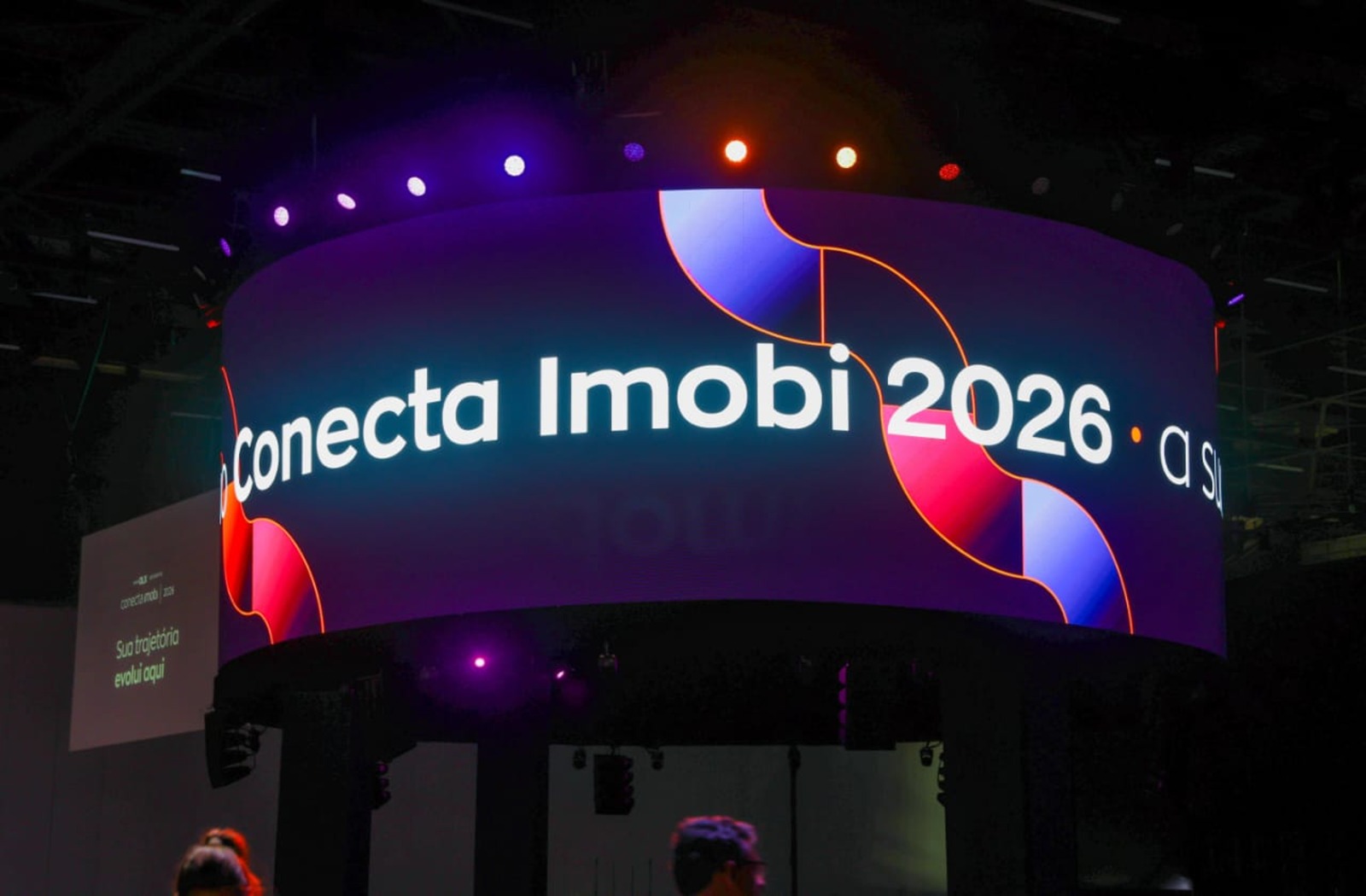 Uma tela LED curva exibe "Conecta Imobi 2026" em branco sobre um fundo abstrato roxo, azul e vermelho em um evento escuro com luzes coloridas.